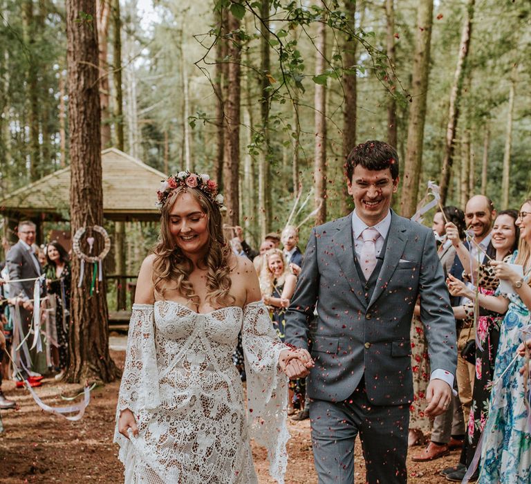 Bride in medieval wedding dress and groom in grey suit with pink tie walks through confetti after handfasting ceremony 