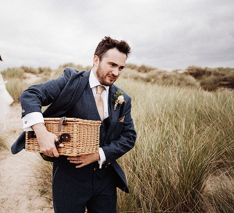 Bride in sheer long sleeve wedding dress carries her shoes along the beach as the groom carries wicker picnic basket