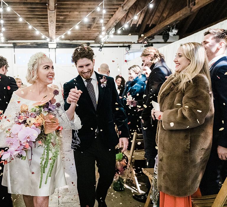 Bride in short wedding dress holding orchid wedding bouquet has confetti exit with groom in velvet suit jacket 