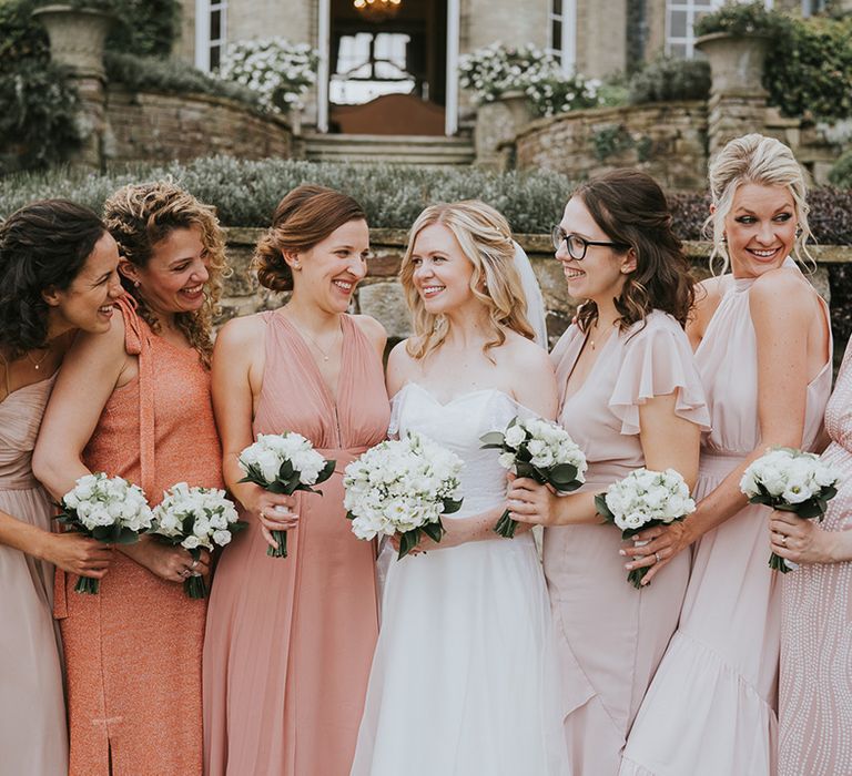 Bridesmaids in pink, peach and coral dresses with bride in off the shoulder wedding dress all holding white rose bouquets 