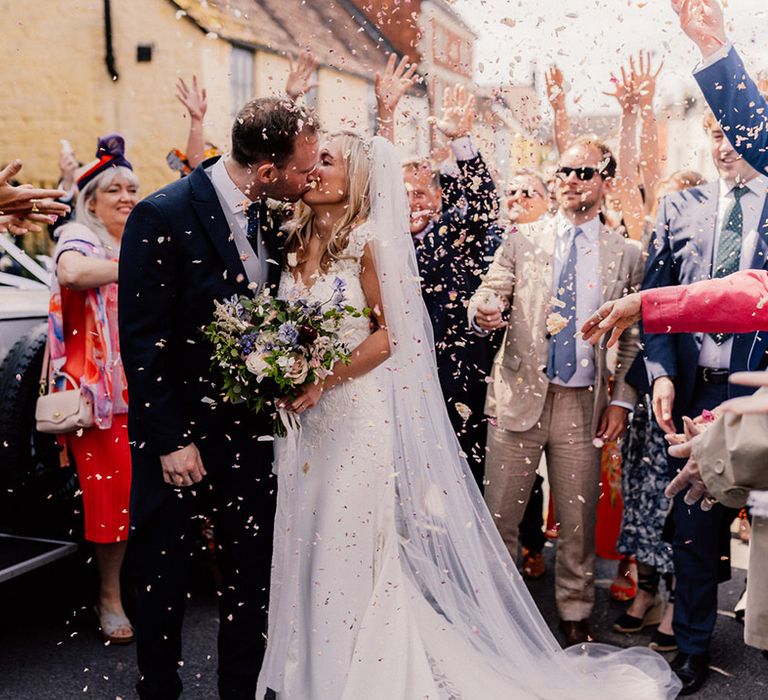 Bride and groom share a kiss outside their black and white wedding car as their guests throw confetti over them 