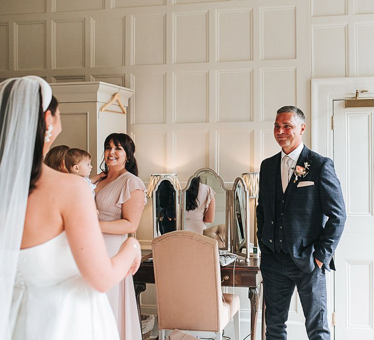 Father of the bride in a dark suit and light tie and blush rose buttonhole smiles as he sees the bride for the first time 