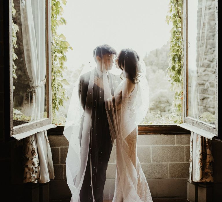 Bride & groom stand beneath pearl embellished veil in front of window on their wedding day