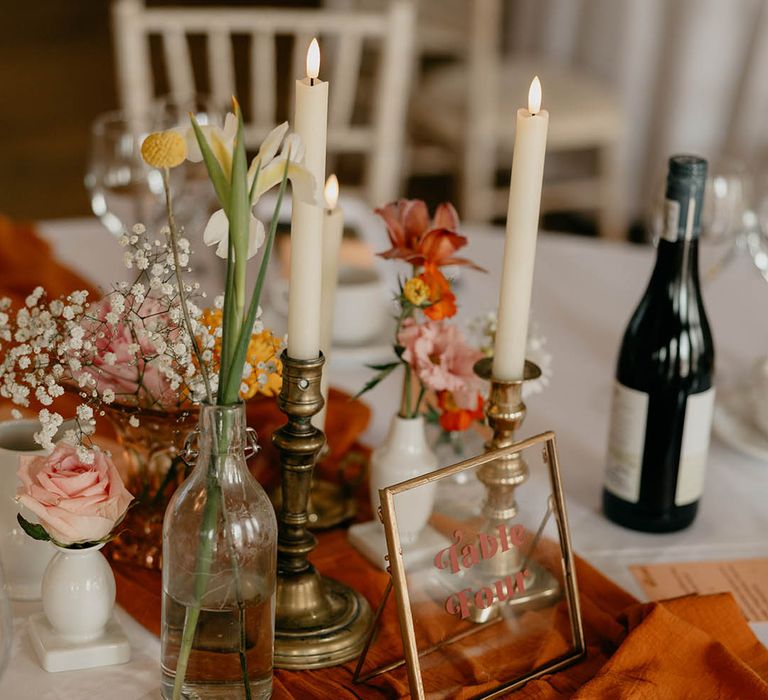 Flower stems, white candles in gold candle holders and mismatched vases decorate the tables with a gold photo frame table number sign