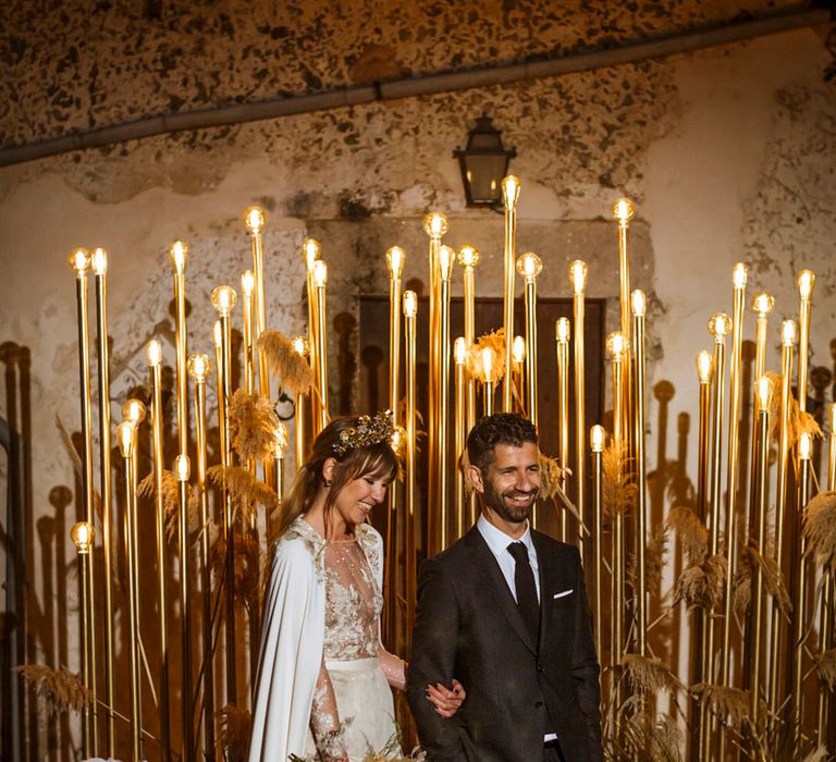 Bride wearing a cape with gold detail and a crown with the groom in a black suit 