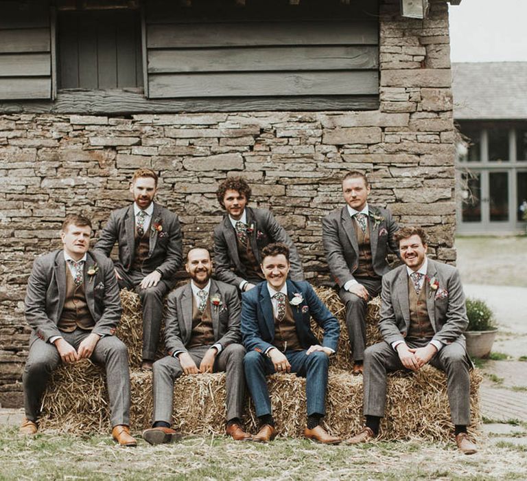 Groom in a blue suit and brown waistcoat with floral tie sitting on a hay bale with the groomsmen in grey suits and brown waistcoats