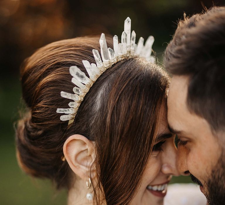 Bride with a pearl necklace, pearl earrings and a quartz crystal crown in a dotted wedding dress rests her forehead against the groom 