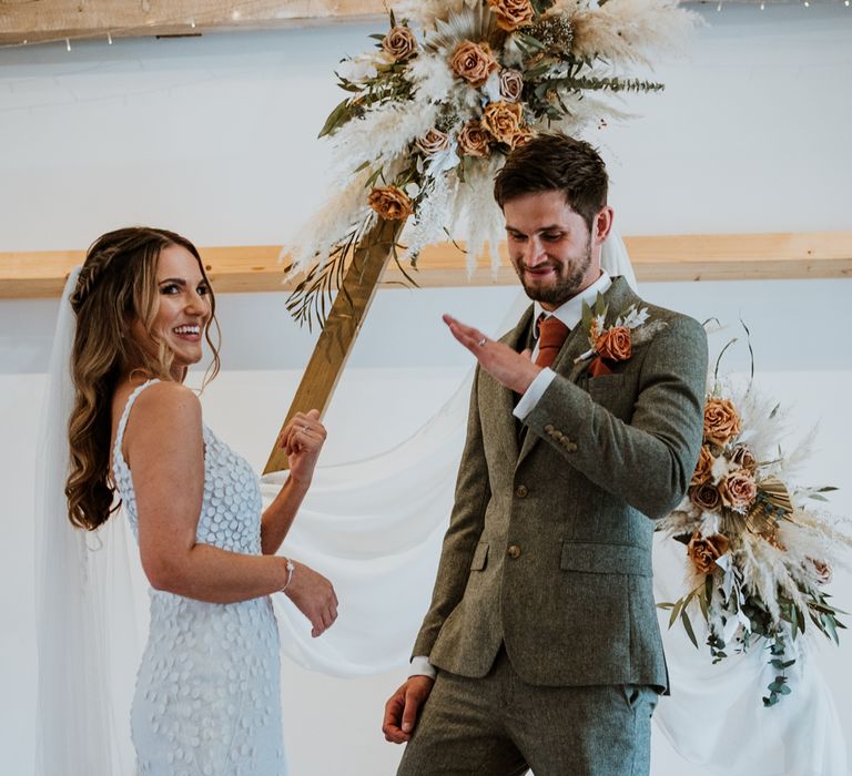 Bride in sleeveless Made With Love Bridal dress with white embellishments and groom in grey suit and pops of burnt orange showing off their new wedding rings at The Green Cornwall wedding venue