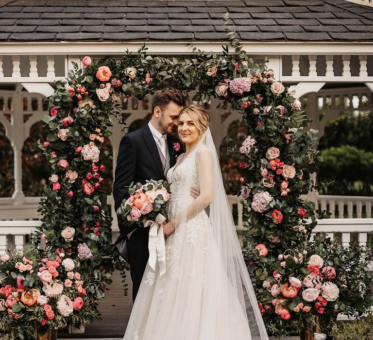 Groom in a dark suit embracing the bride in a lace wedding dress in front of the pink flower arch altar decor 