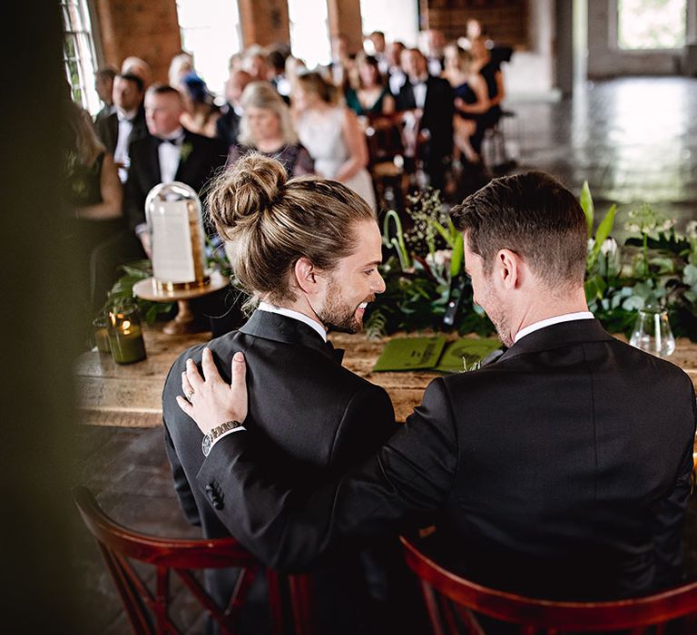 Grooms sit at wooden rustic table during botanical styled wedding ceremony in industrial setting