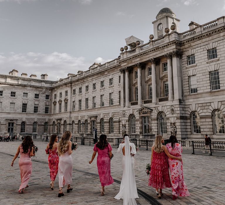 Bride in modern wedding dress with open back detail and long veil walking with bridal party wearing mismatched pink bridesmaid dresses at Somerset House