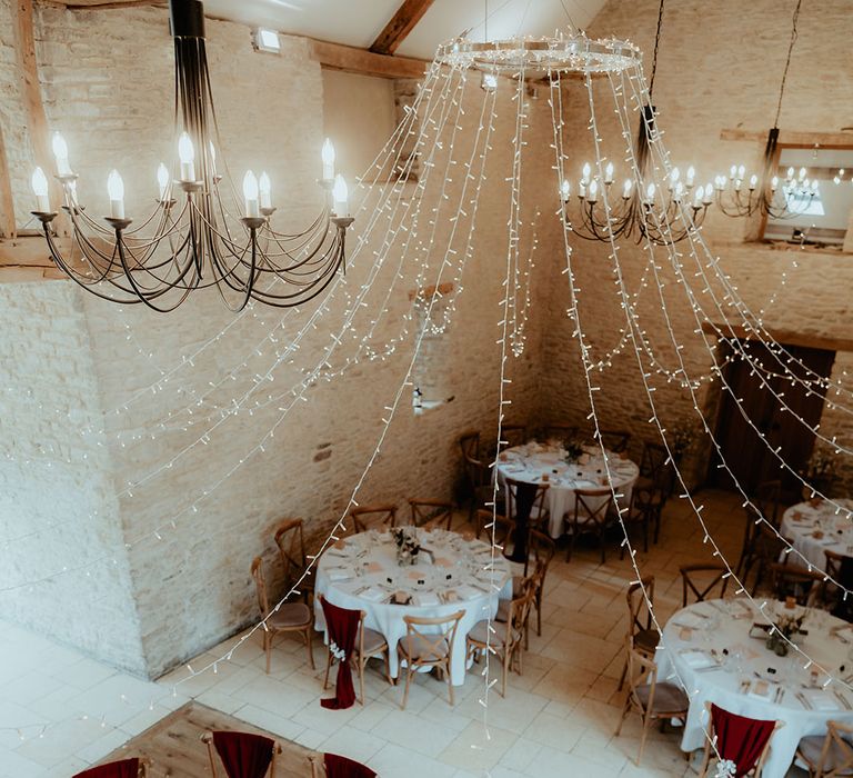 Reception room decorated with fairy lights, round tables and gold touches with burgundy chair back decor and rustic details 