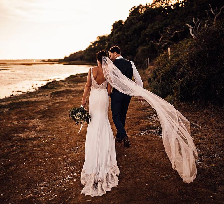 The bride and groom walk along the beach with the bride's veil blowing in the wind