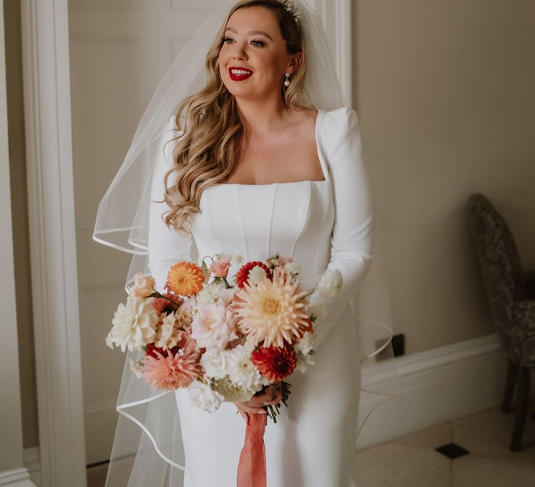 Bride in square neck long sleeve satin wedding dress with two tier veil, pearl earrings and bridal crown holding mixed colourful floral bouquet