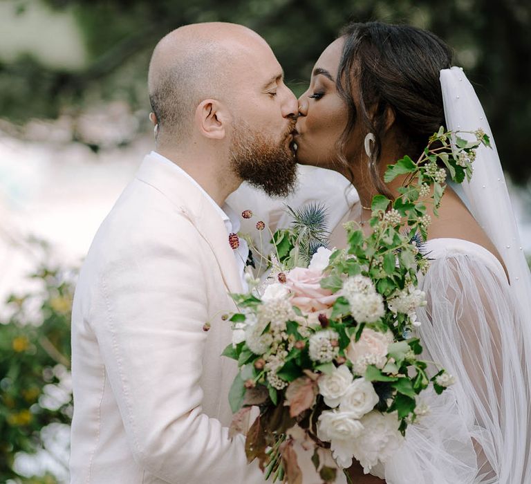 Bride and Groom kiss with bouquet of Rosses, thistles and wildflowers in the foreground