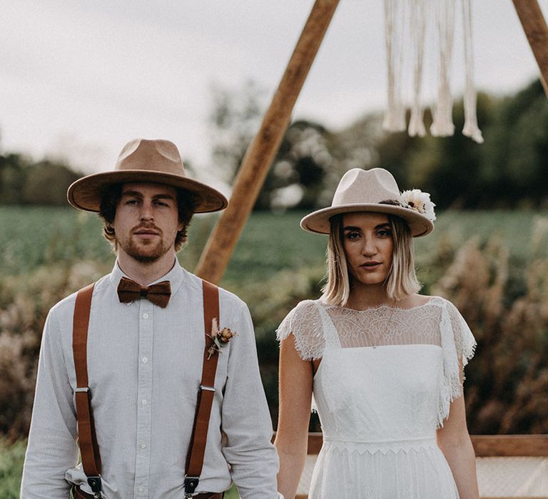 The bride and groom pose together and look into the camera both wearing their stylish hats 