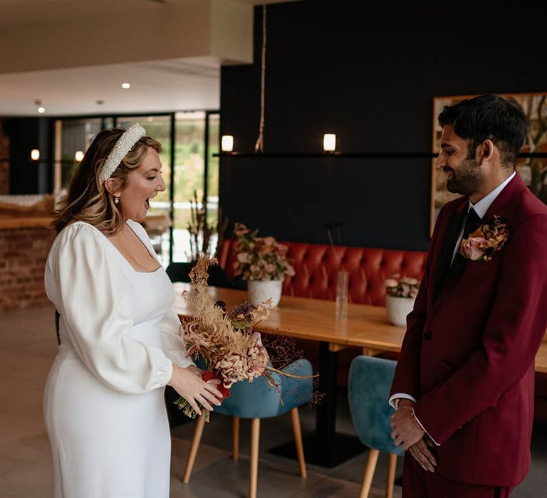 Groom in burgundy suit smiles as he sees the bride for the first time and the bride smiles brightly as she sees the groom for their first look moment