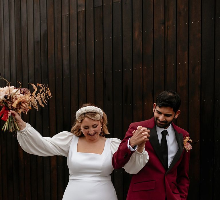 Bride with pearl headband and long sleeve square neckline wedding dress holding neutral bouquet walking with the groom in burgundy suit jacket and black details