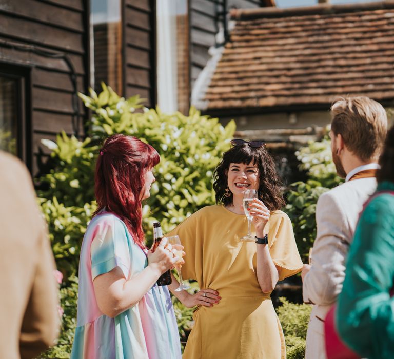 Wedding guest wearing mustard coloured boxy dress having a glass of champagne and chatting with other guests