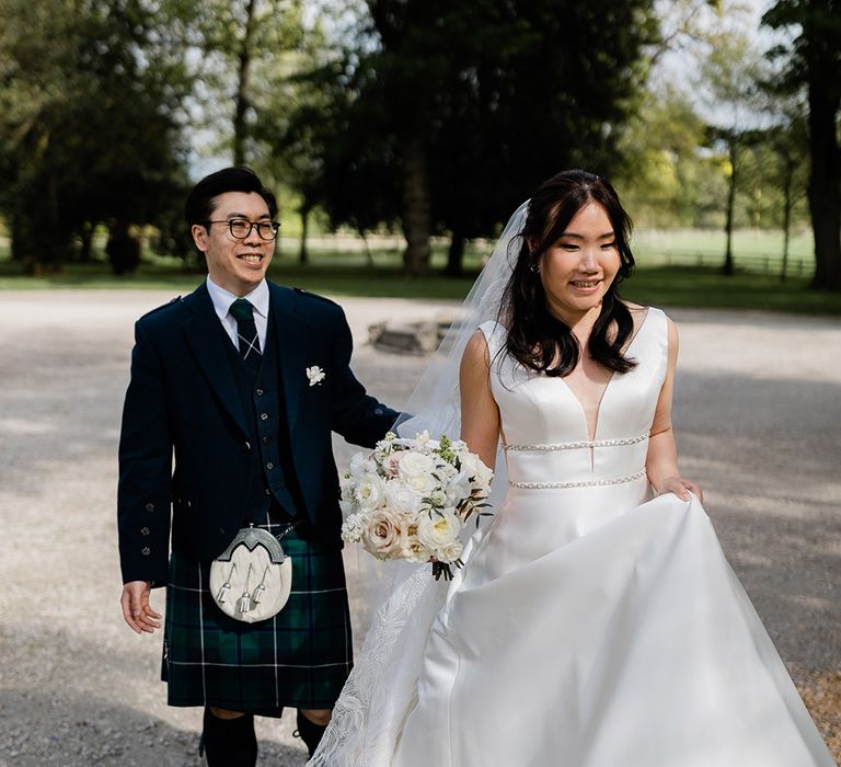 Bride wears Princess wedding dress with embellished belt to waistband and walks alongside her groom in green kilt