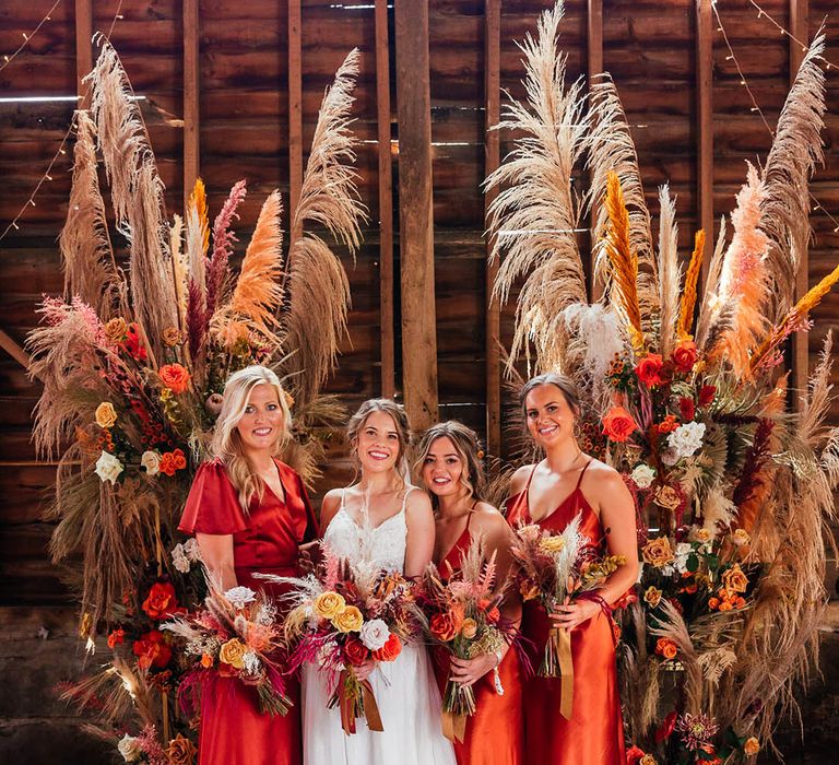 Bridesmaids in satin burnt orange dresses with the bride in a lace wedding dress in front of pampas grass boho flower columns with roses 