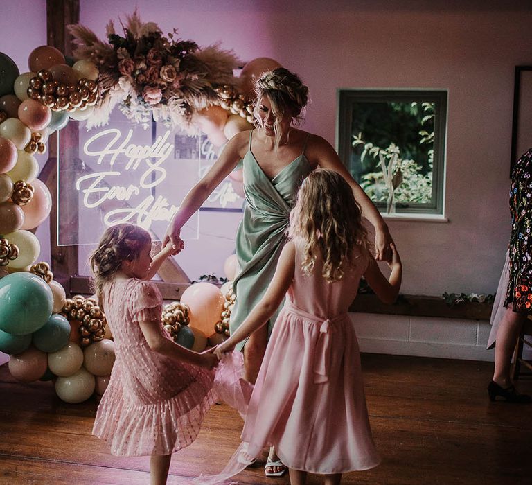 Bridesmaid in a sage green satin dress dancing with flower girls in pink dresses next to the neon sign and balloon arch decor 