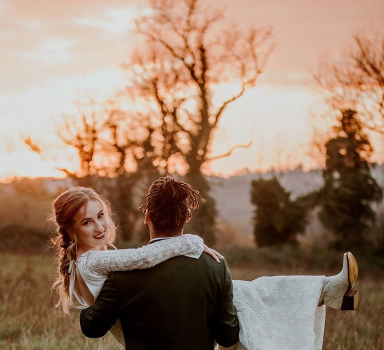 Bride in boho long sleeve lace wedding dress with tassels with open back detail being carried by groom in black tux 