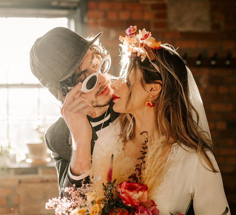 Bride in three quarter length sheer sleeve wedding dress with button and cinched waist details and pink bridal flower crown embracing groom in black and white long sleeve shirt, cap and large sunglasses at jumping the broom wedding ceremony 