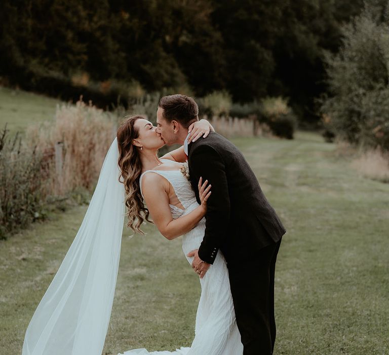 Groom in black suit kiss the bride as he dips her for cute couple portraits for the rustic glam wedding at Rhyse Farm 