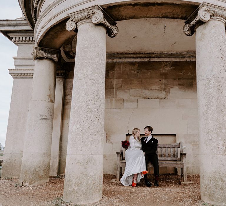 The bride in a metallic wedding dress wearing bright orange heels sitting on a bench with the groom in a pinstriped suit 