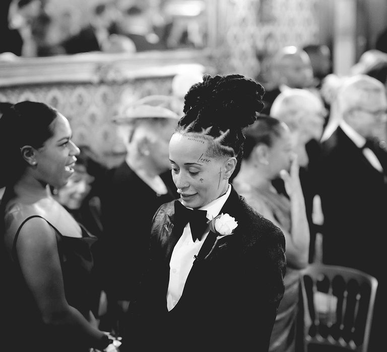 Bride in black-tie tuxedo with black bowtie, white rose boutonniere waiting at the end of the aisle at Grittleton House