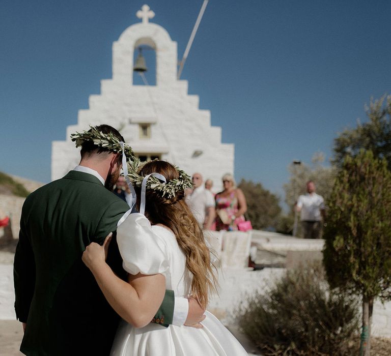 Bride and Groom embracing and wearing Stefana crowns in front of white greek chapel at destination wedding in Paros