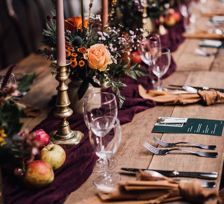 Purple and peach taper candle in gold candleholders with dark red table runner, dark green wedding menu, and mustard yellow napkins for autumnal wedding tablescape 