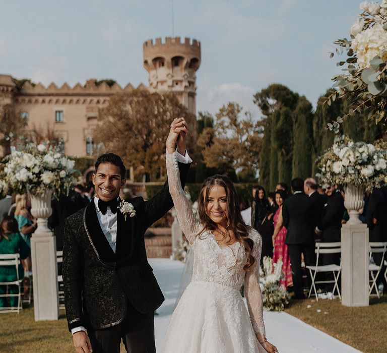 Groom in classic black tuxedo with white flower boutonniere posing with bride in lace long sleeve Pronovias wedding dress and church-length veil at castle wedding venue Barcelona  