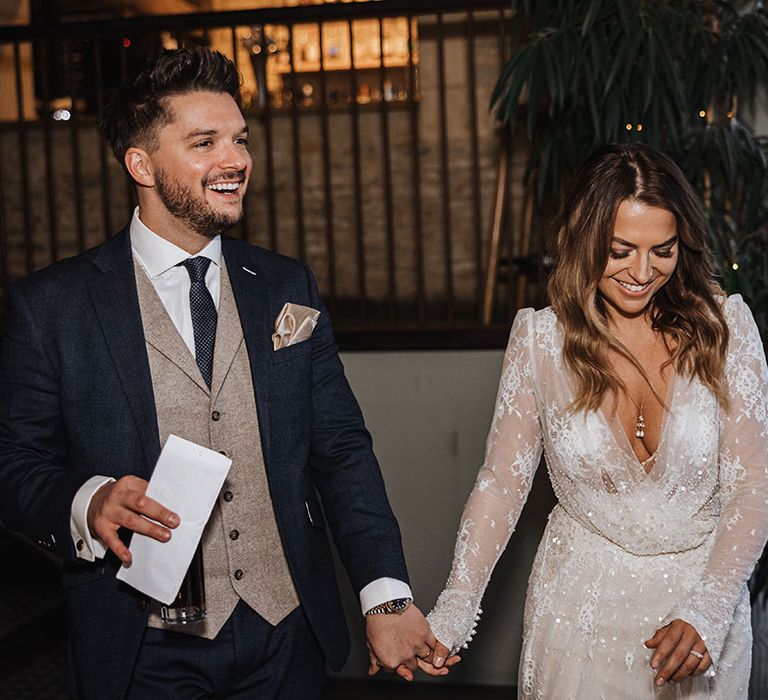 Groom in navy suit with a cream waistcoat walking into the wedding reception with the bride in a long sleeve sparkly winter wedding dress