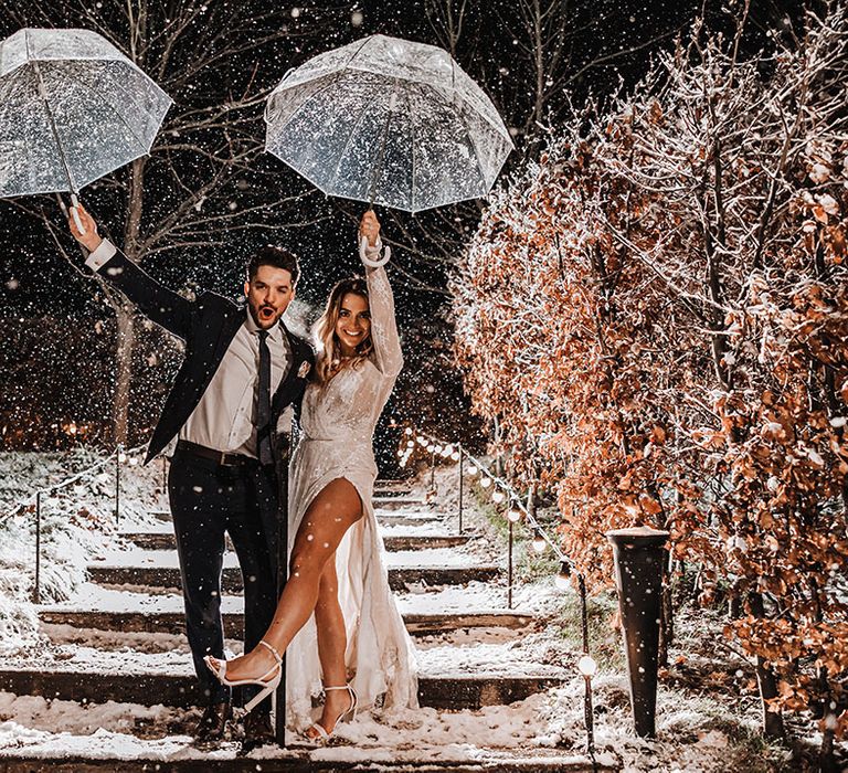 Bride in long sleeve sparkly wedding dress with white block heels raising her umbrella in the air with the groom doing the same for cute winter wedding couple portraits 