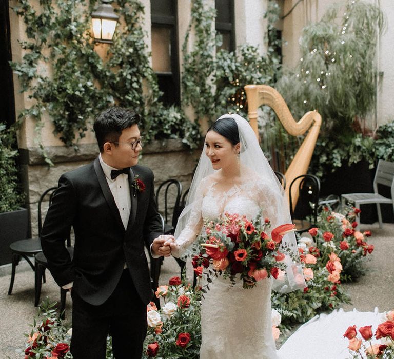 Bride in long sleeve lace wedding dress and veil carrying red flower bouquet walking down the aisle to meet the groom for their private first look