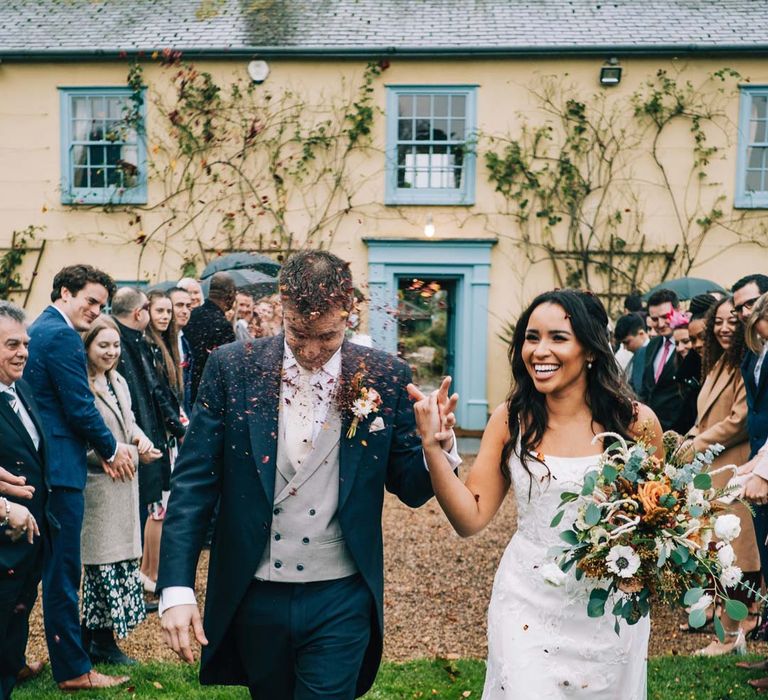 Bride in a-line tulle wedding dress with beaded lace applique with petals and a square neckline with side tulle cutouts holding pampas grass, eucalyptus, blush garden roses, dried flowers and foliage bridal bouquet walking with groom in navy grooms morning suit with grey waistcoat, off-white tie and mixed wildflower boutonniere doing dried flower autumnal confetti exit at South Farm wedding 