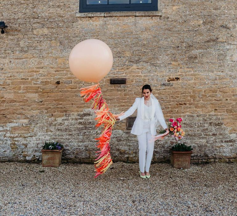 Bride in white bridal suit with closed toe bright green wedding heels holding large balloon with wedding streamers attached outside of Merriscourt wedding venue