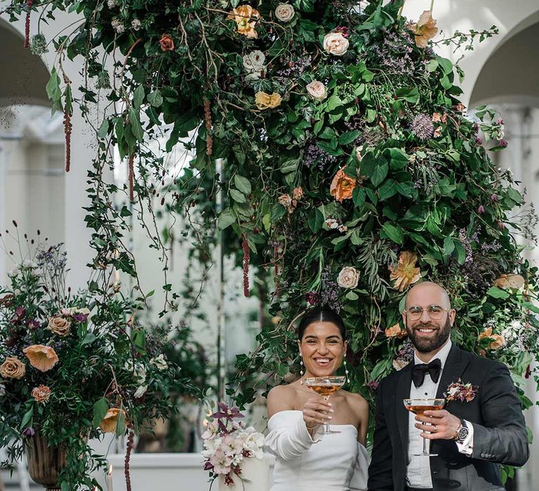 Bride in long sleeve off the shoulder wedding dress with pearl button detailing and a side slit standing with groom in classic black tuxedo with bowtie and wildflower boutonniere holding up cocktails standing by dessert table surrounded by suspended wildflower and foliage arrangements in large gold vases at Buxted Park