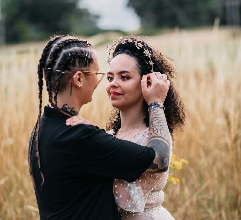 Bride in silver chains, glasses, black shirt and black trousers dancing with bride in fields wearing long sleeve v-neck sheer polka dot overlay sparkly wedding dress and gold celestial bridal headband 