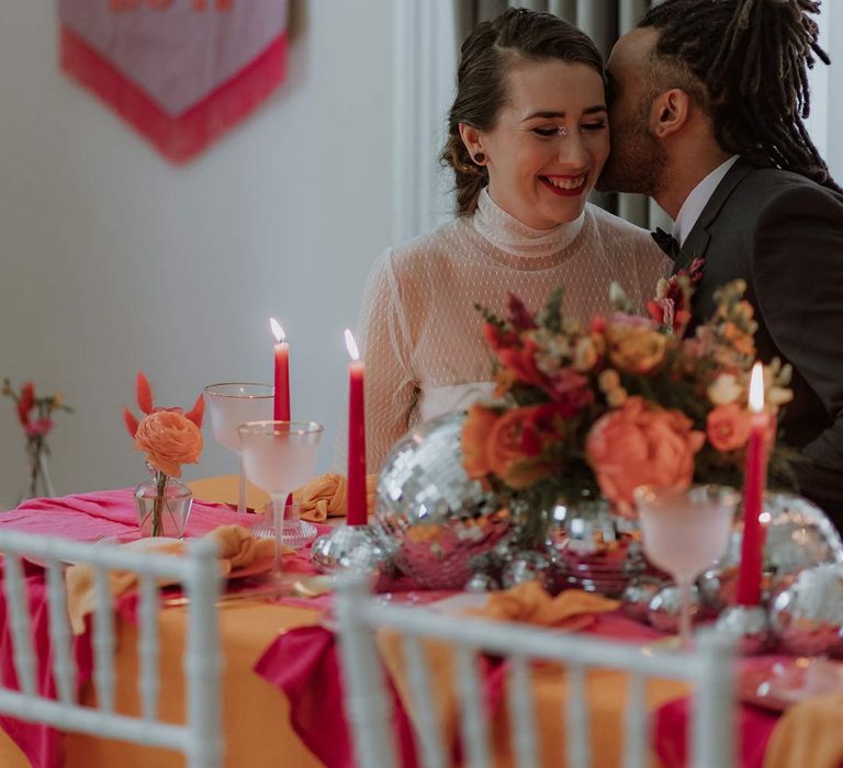 Groom in black tuxedo embracing bride in mesh long sleeve high neck polka dot wedding dress sat at pink and orange wedding tablescape with disco ball decor and bright floral centrepieces 