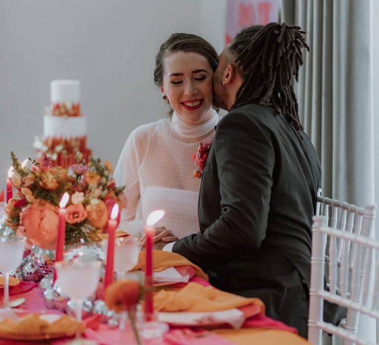 Bride in mesh long sleeve high neck polka dot wedding dress with groom in black suit sat at pink and orange wedding tablescape with orange tablecloth, bright pink wedding table runners, orange wedding napkins, pink tapered candles, pink and orange poppies, garden roses, carnations, dried flowers and foliage wedding table centrepieces and disco ball wedding decor 