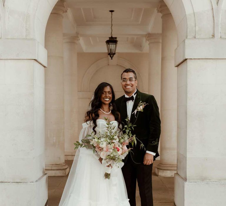 Bride in off the shoulder layered tulle wedding dress and floral embroidered cathedral-length veil holding large mixed bridal bouquet standing with groom in bottle green velvet grooms suit, black bowtie and chic boutonniere 