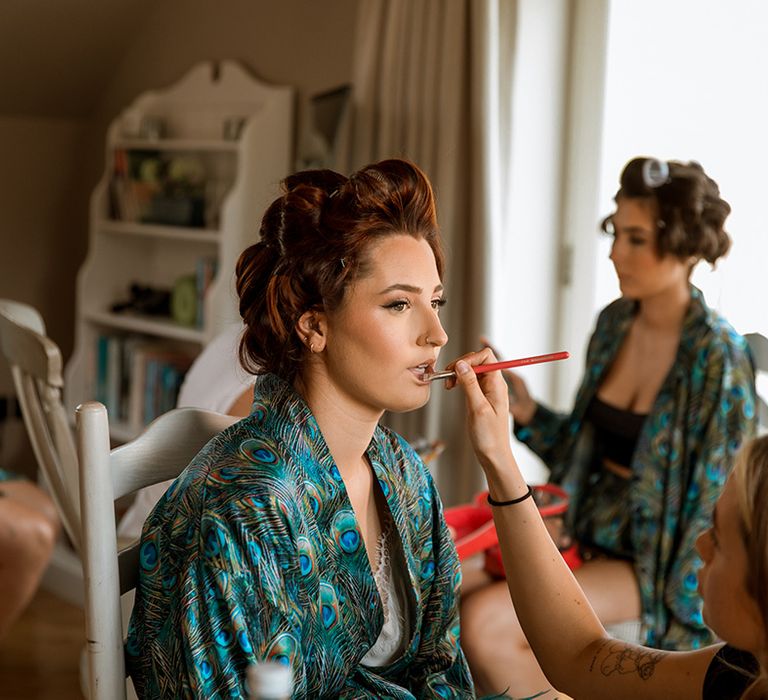 Bride in peacock feather satin robe getting her makeup done for the wedding day 