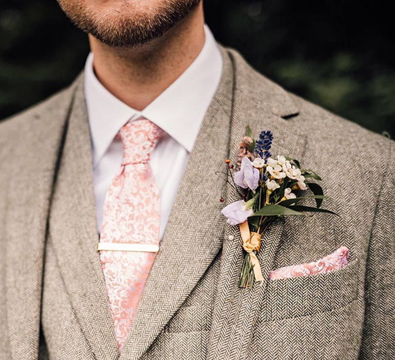 Groom in three piece tweed grey suit with patterned pink tie and floral buttonhole 