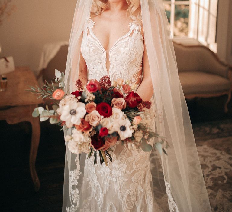 Bride wearing embellished lace wedding dress with embroidered veil holding white anemone and red rose bouquet 