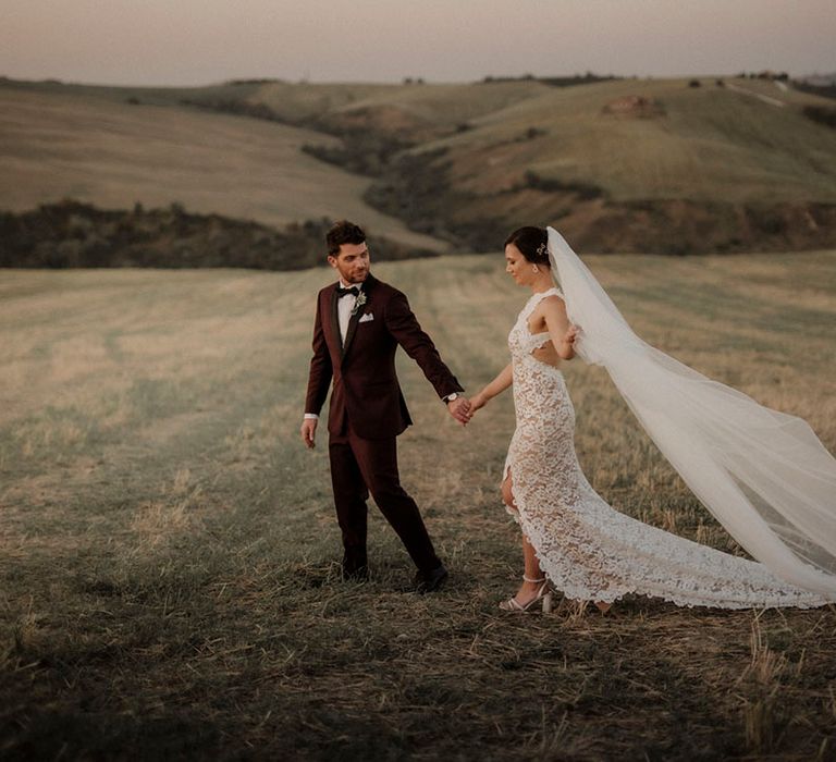 Bride in stunning lace bridal gown with long cathedral length veil with groom in burgundy suit 