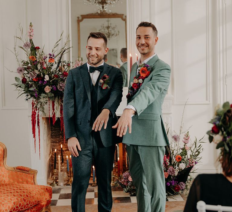 Groom in dark blue wedding suits with groom in mint green wedding suit standing together as they show off their new wedding bands 