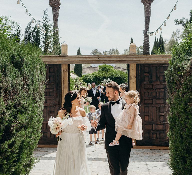 Bride and groom walking with their daughter carrying champagne glasses to celebrate the wedding 